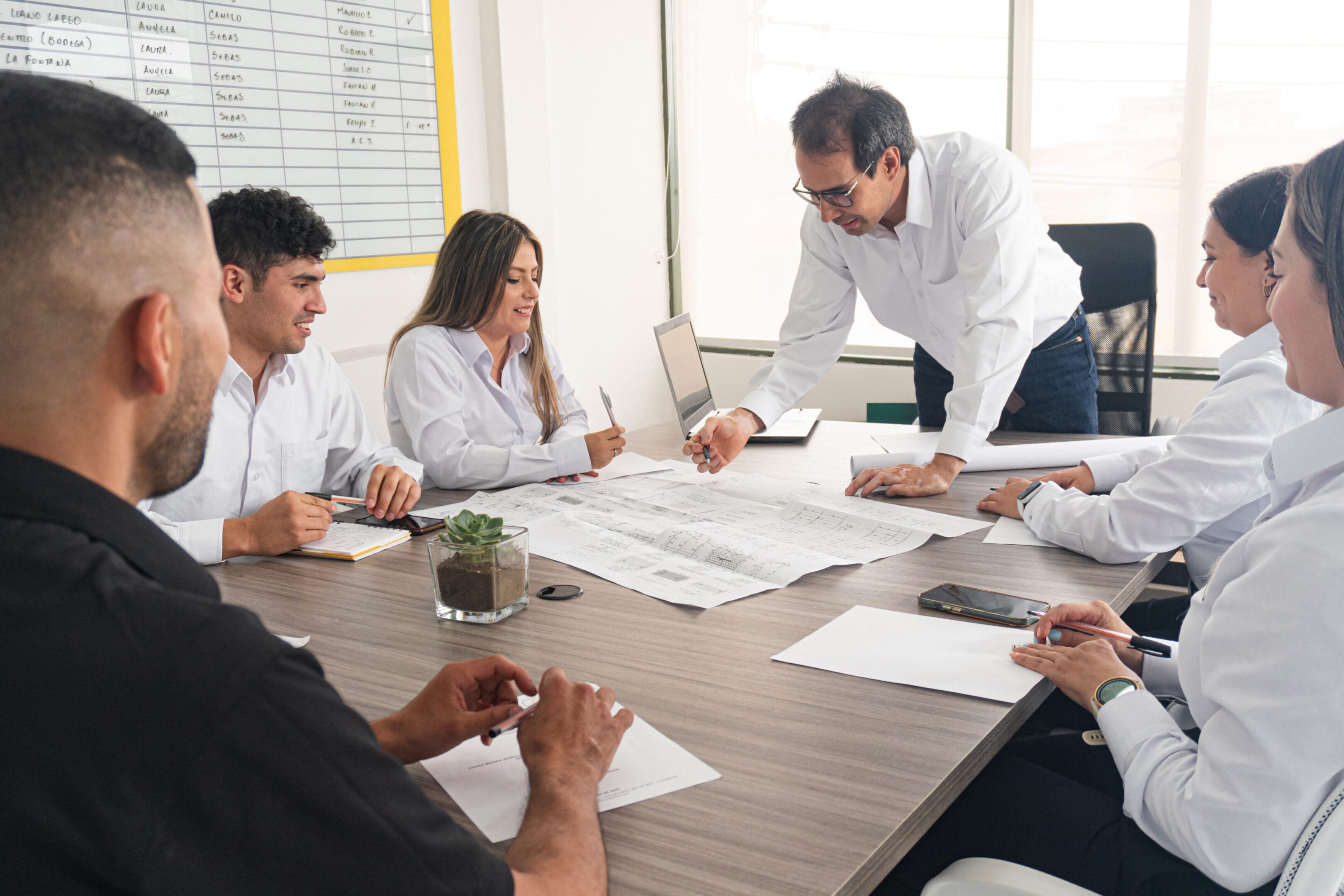Person with vitiligo in a work environment. Business meeting and project planning at an engineering company. A business meeting at an engineering firm, where professionals discuss projects and delivery dates. Participants review blueprints, take notes, and analyze a whiteboard to make strategic decisions. The atmosphere is collaborative and focused on achieving set goals.